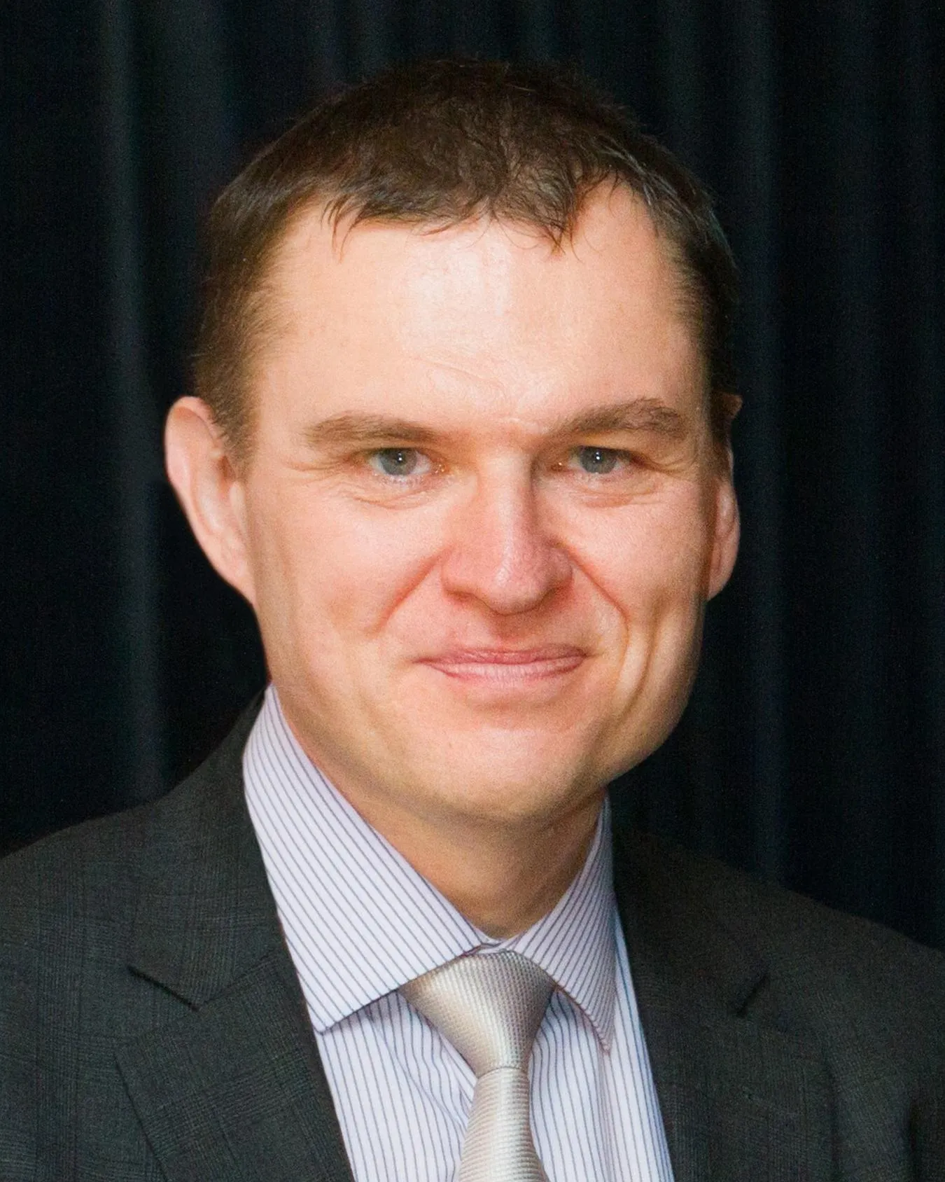 Portret mężczyzny w garniturze z uśmiechem na ciemnym tle.