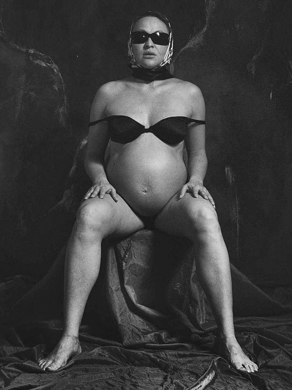 Kobieta w ciąży w ciemnych okularach z chustą na głowie. Ma na sobie tylko stanik i siedzi w lekkim rozkroku na przykrytym materiałem stołku