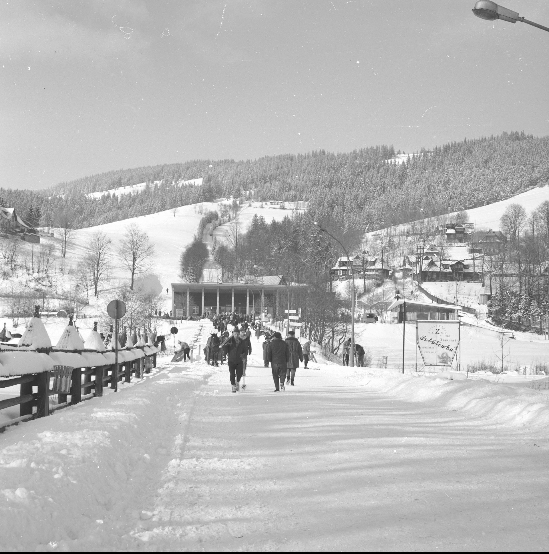 Turyści na deptaku pod Gubałówką. W tle dolna stacja kolejki linowo-terenowej i pasmo Gubałówki.